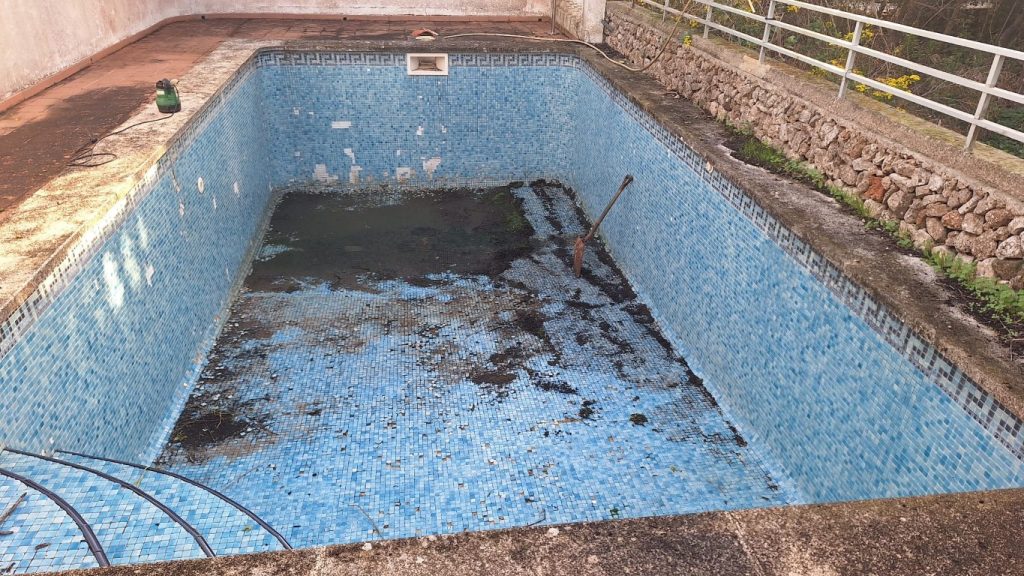 Piscina rectangular de mosaico azul en Menorca, con azulejos visibles y agua estancada, borde de piedra y baranda, lista para mantenimiento.