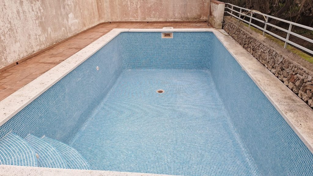 Piscina rectangular de mosaico azul claro vacía, borde de piedra arenosa, escaleras curvas y desagüe central, ubicada en Menorca, lista para mantenimiento y reformas.